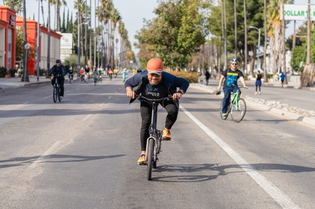 Man in dark pants and long-sleeved shirt, wearing an orange helmet, leaning over his bike as he cycles down a wide street, with other cyclists in the background behind him.
